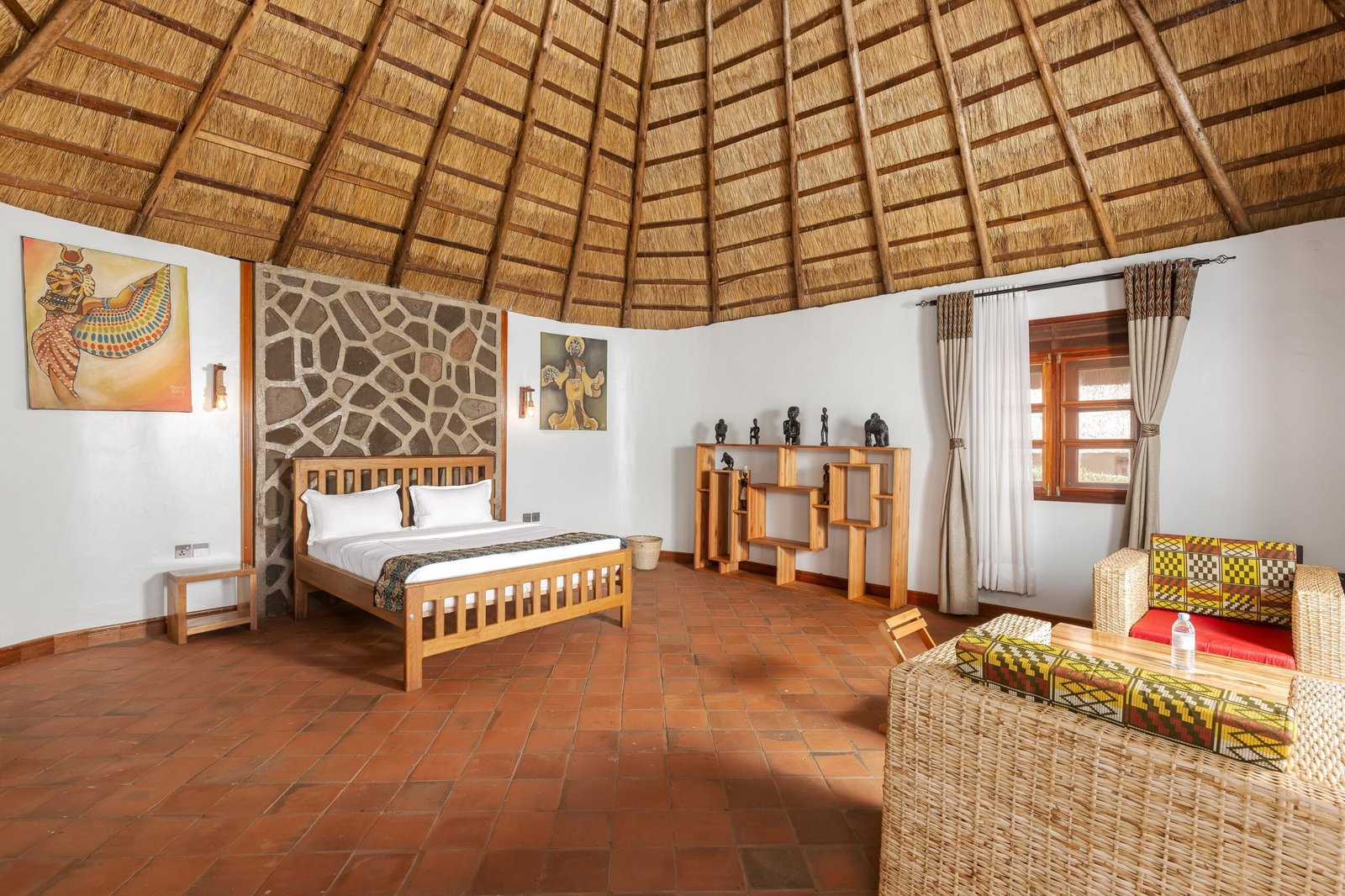 A king cottage bedroom beneath the round thatched roof, with a stone feature wall and local artwork