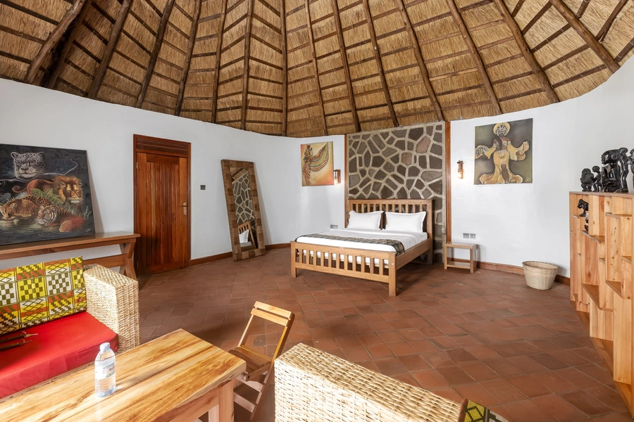 A king cottage interior at Bahundu, with a king bed beneath the round thatched roof and local artwork on the walls, near Kibale Forest in western Uganda